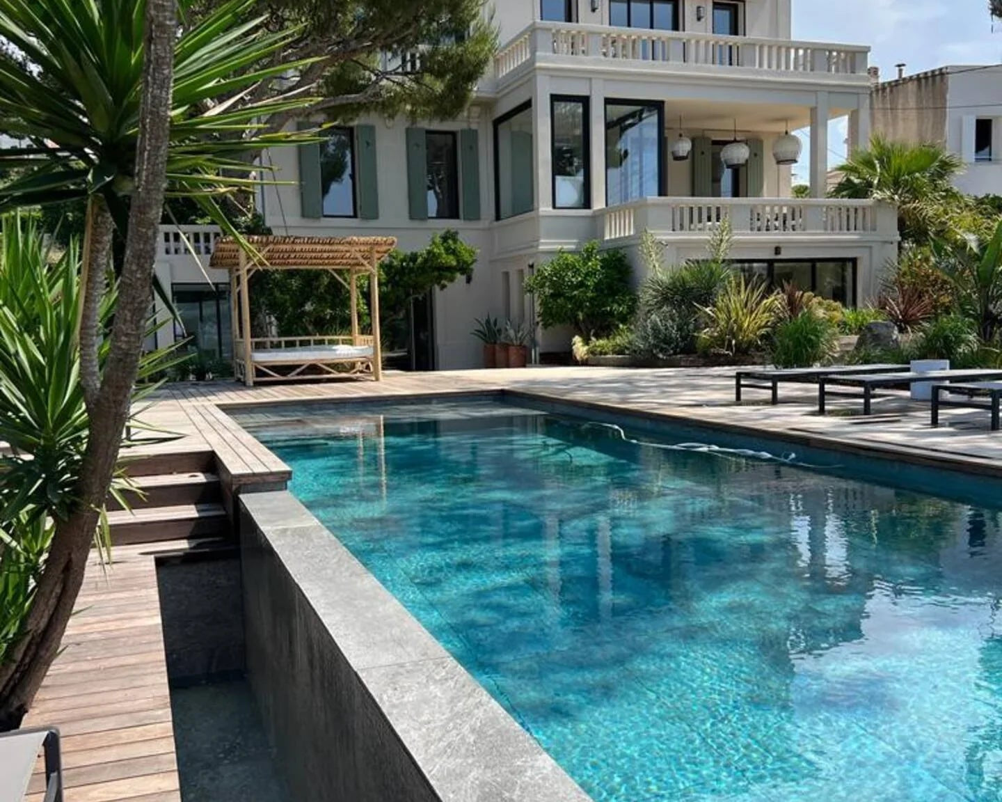 Piscine moderne et maison blanche avec terrasse et jardin méditerranéen sous un ciel bleu