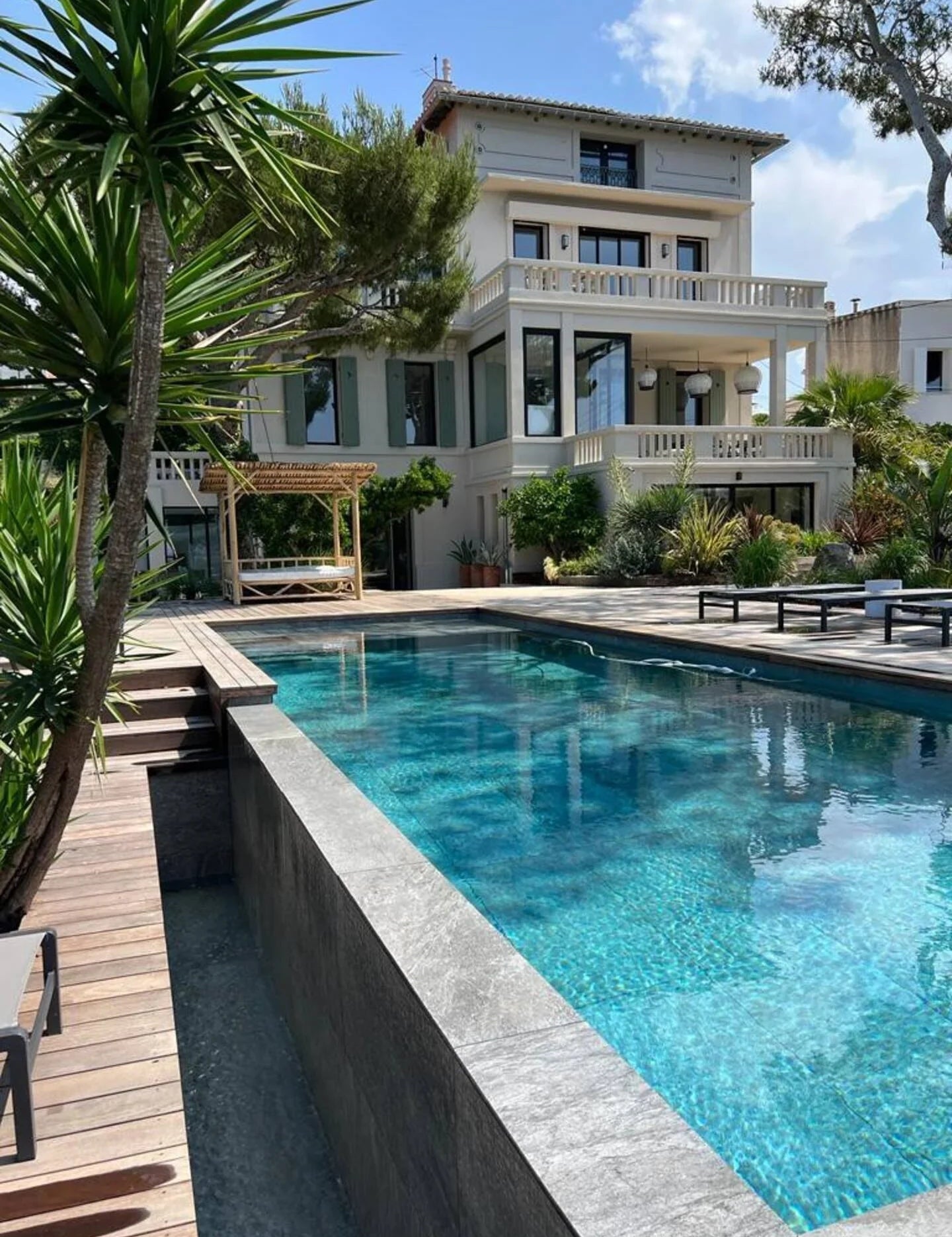 Piscine moderne et maison blanche avec terrasse et jardin méditerranéen sous un ciel bleu