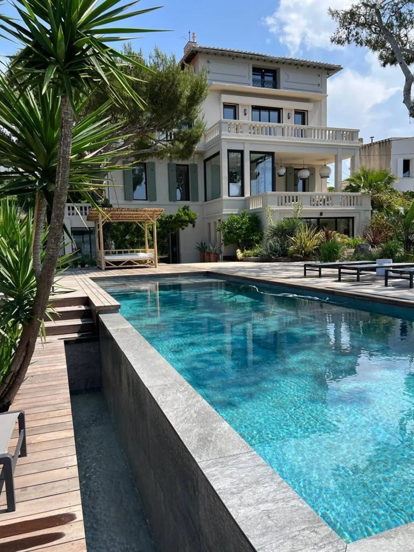 Piscine moderne et maison blanche avec terrasse et jardin méditerranéen sous un ciel bleu