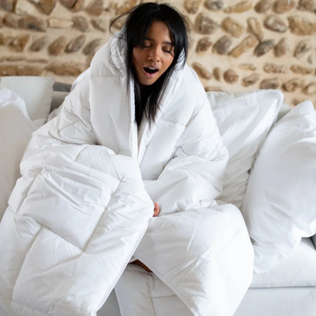 Jeune femme enveloppée dans une couette blanche moelleuse assise sur un canapé avec mur en pierre