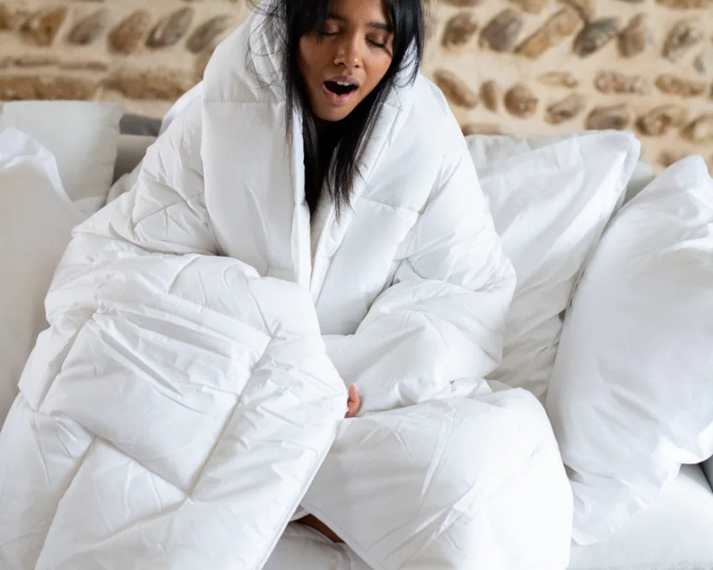 Jeune femme enveloppée dans une couette blanche moelleuse assise sur un canapé avec mur en pierre