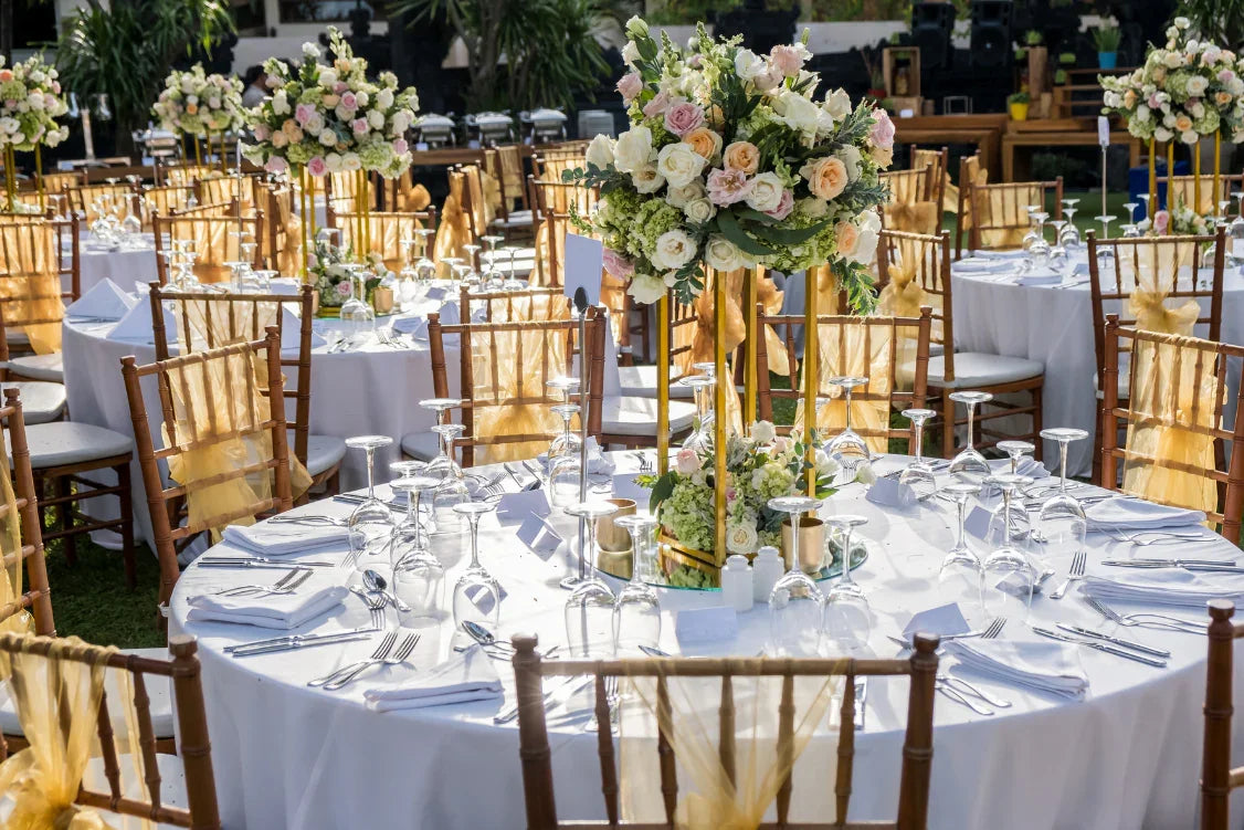 Table de réception élégante avec nappes blanches, chaises bois décorées de voilages dorés et centres de table floraux