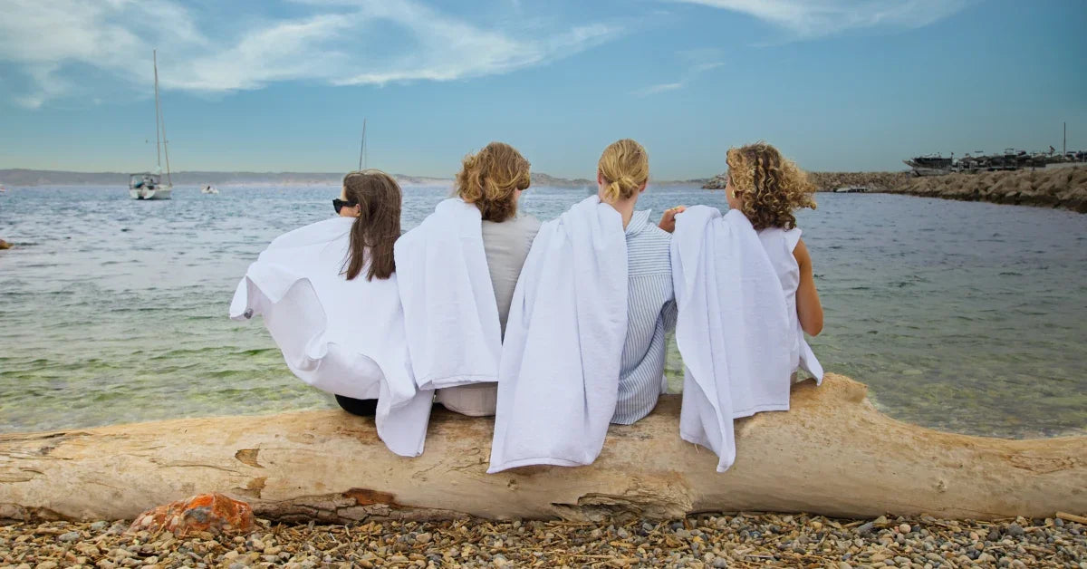 Quatre femmes assises sur un tronc au bord de la mer, couvertes de serviettes blanches, devant des voiliers