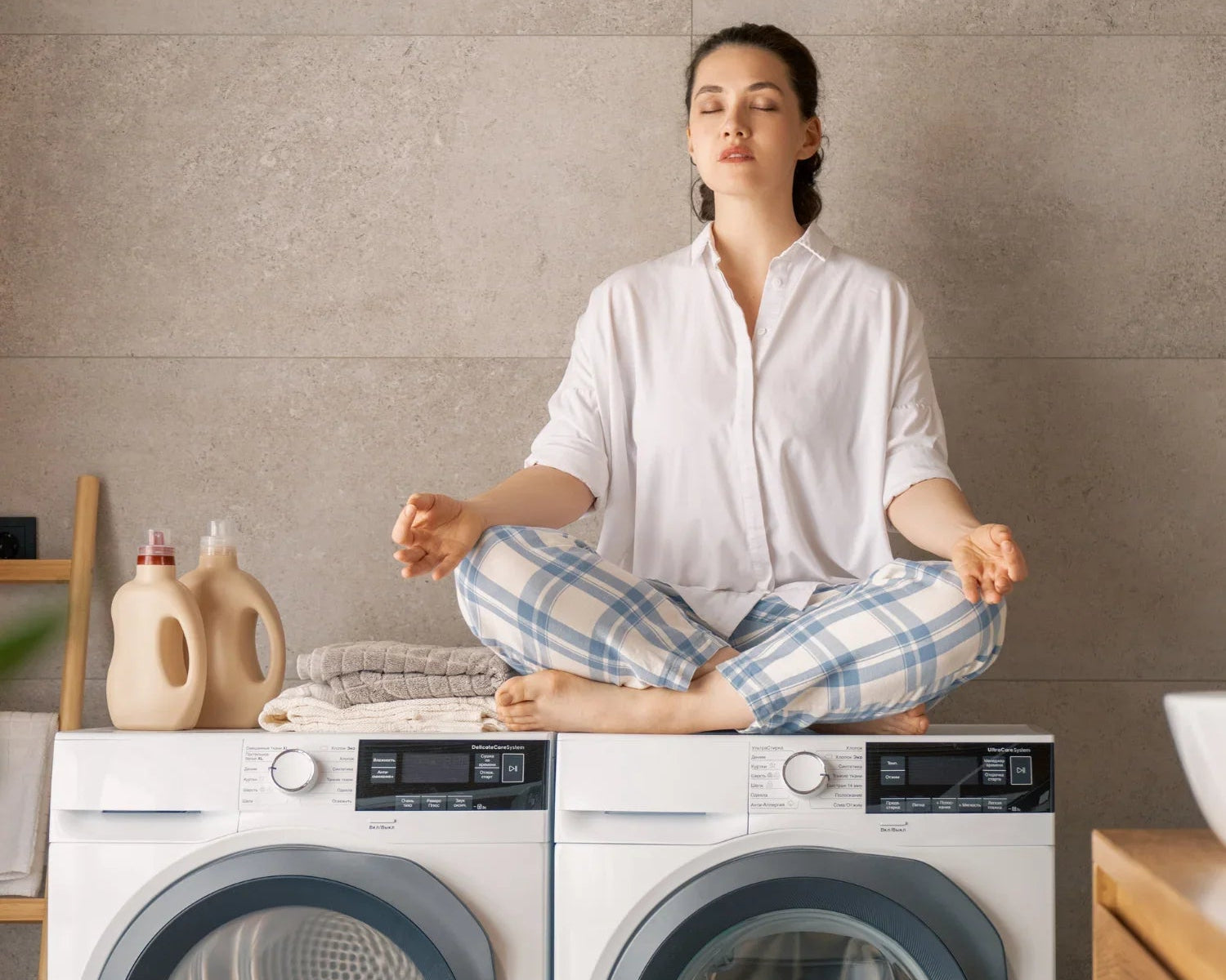 Femme méditant en pyjama sur machine à laver avec détergents et serviettes pliées dans buanderie