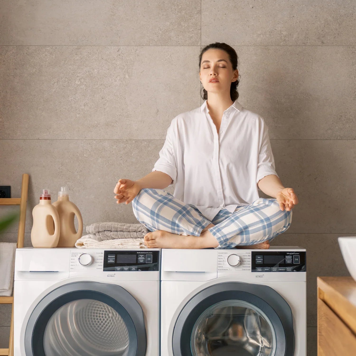 Femme méditant en pyjama sur machine à laver avec détergents et serviettes pliées dans buanderie