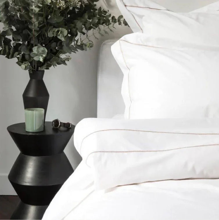 Chambre avec draps blancs élégants et couture beige, table noire avec vase et plante verte
