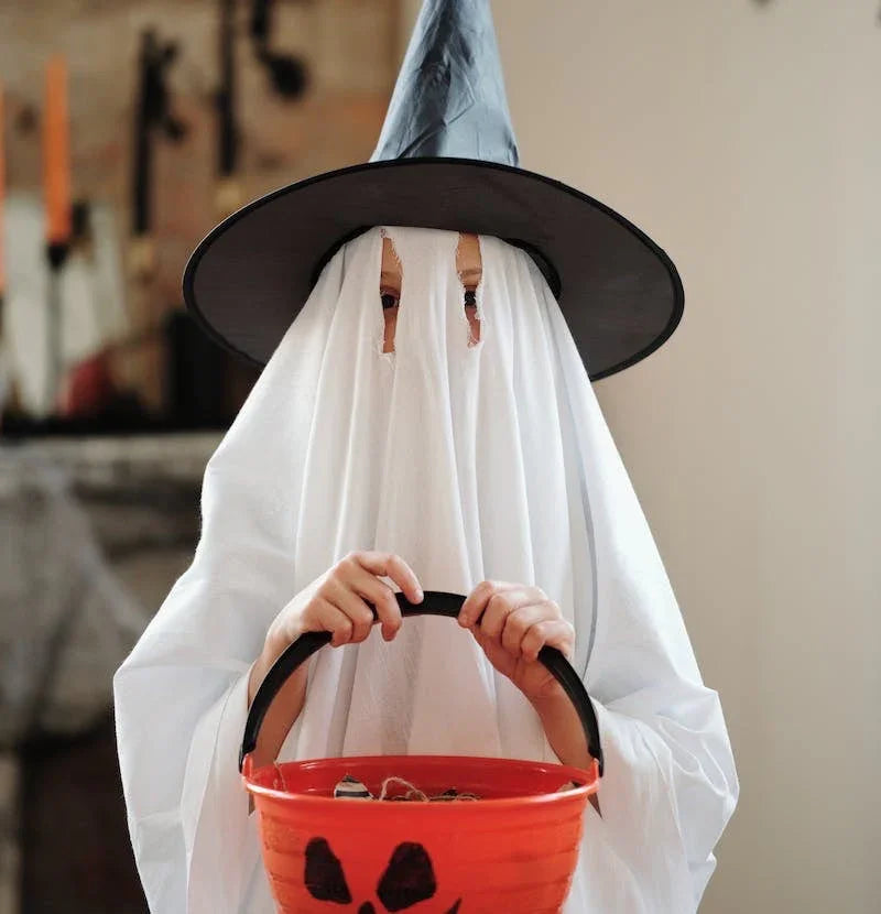 Enfant déguisé en fantôme avec drap blanc et chapeau de sorcière tenant un seau orange Halloween