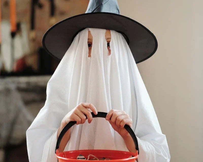 Enfant déguisé en fantôme avec drap blanc et chapeau de sorcière tenant un seau orange Halloween