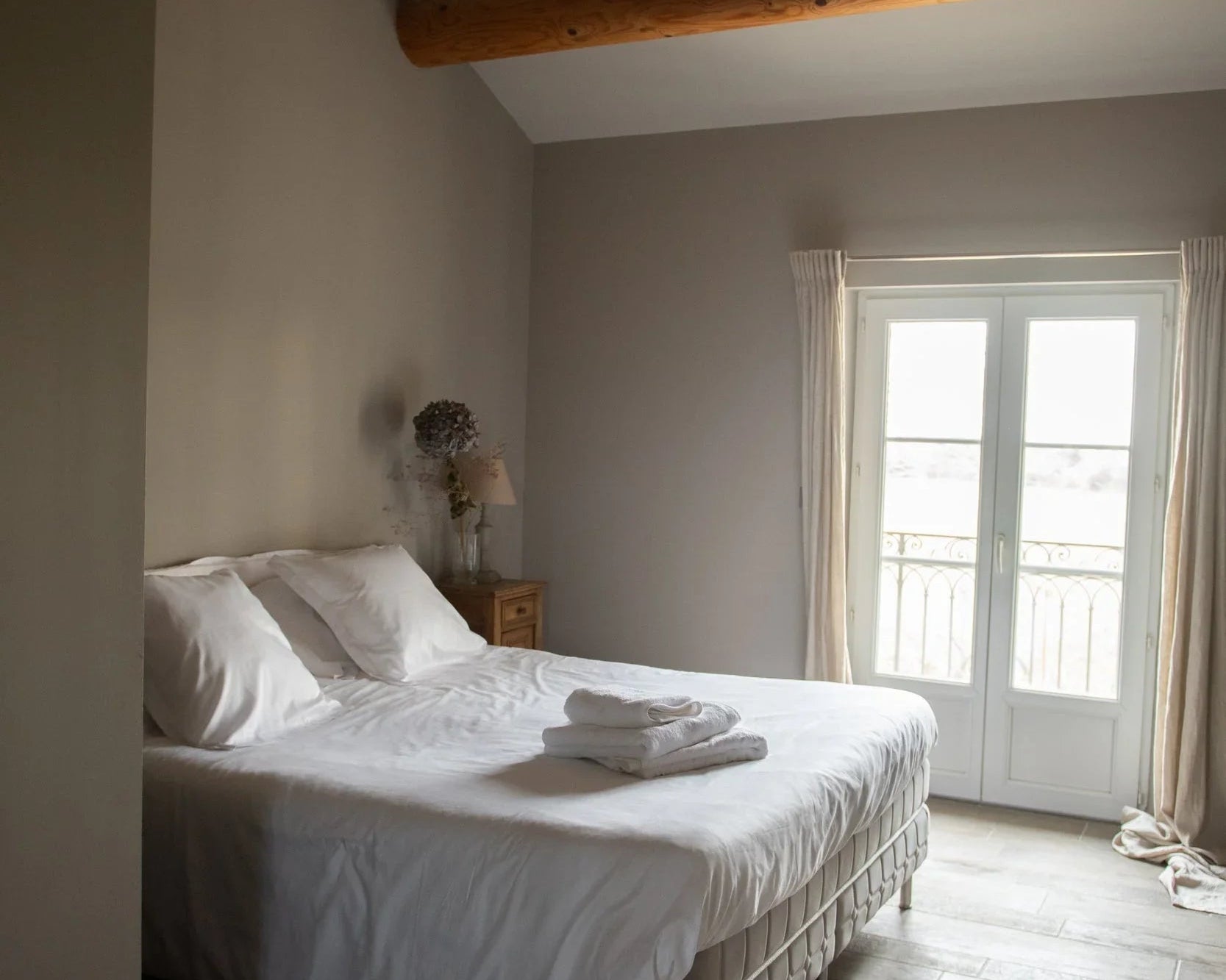 Chambre lumineuse avec lit double, draps blancs, poutres bois au plafond, porte-fenêtre lumineuse