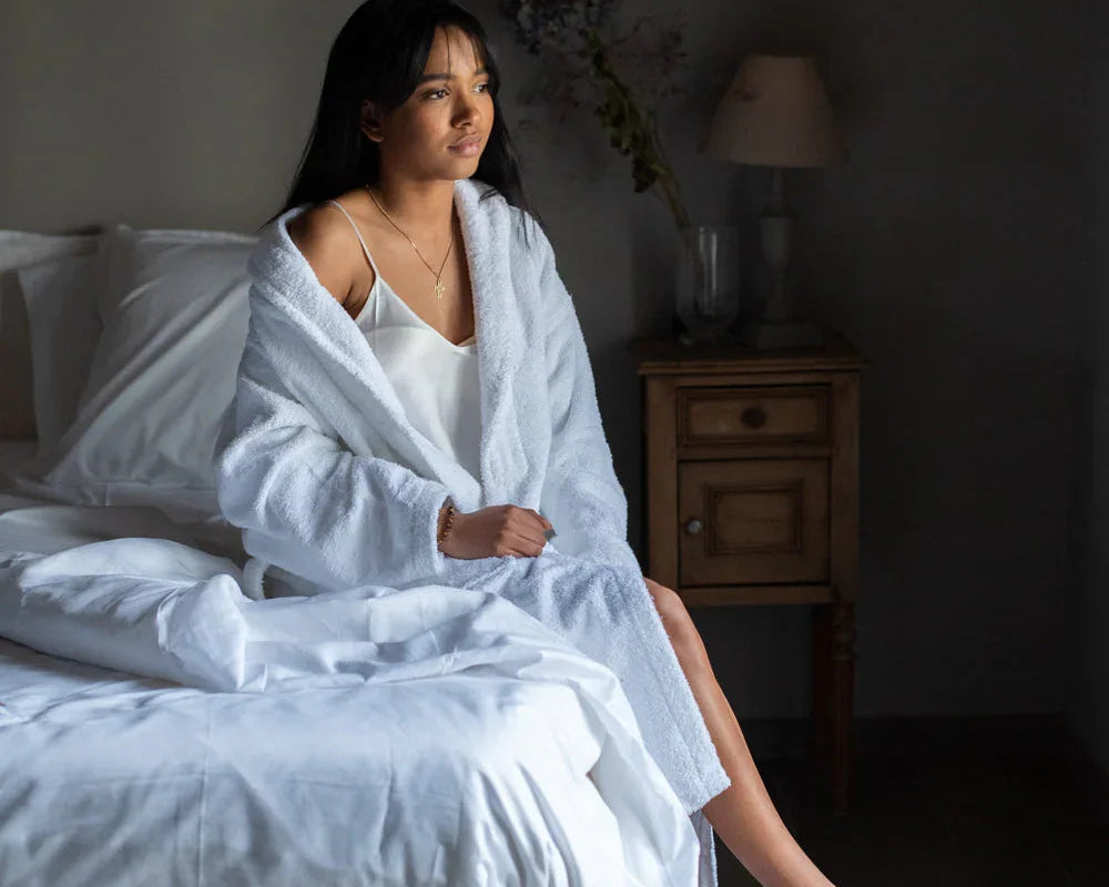 Femme en peignoir blanc et chaussons sur le bord d'un lit dans une chambre minimaliste