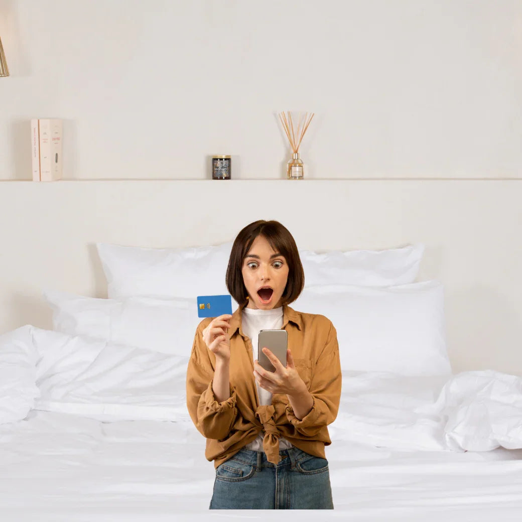 Femme surprise avec carte bancaire et smartphone devant lit avec linge hôtelier blanc