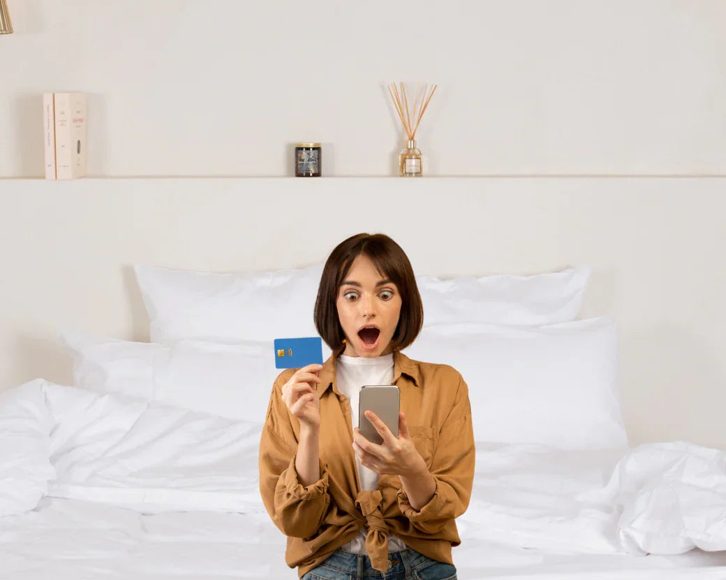 Femme surprise avec carte bancaire et smartphone devant lit avec linge hôtelier blanc
