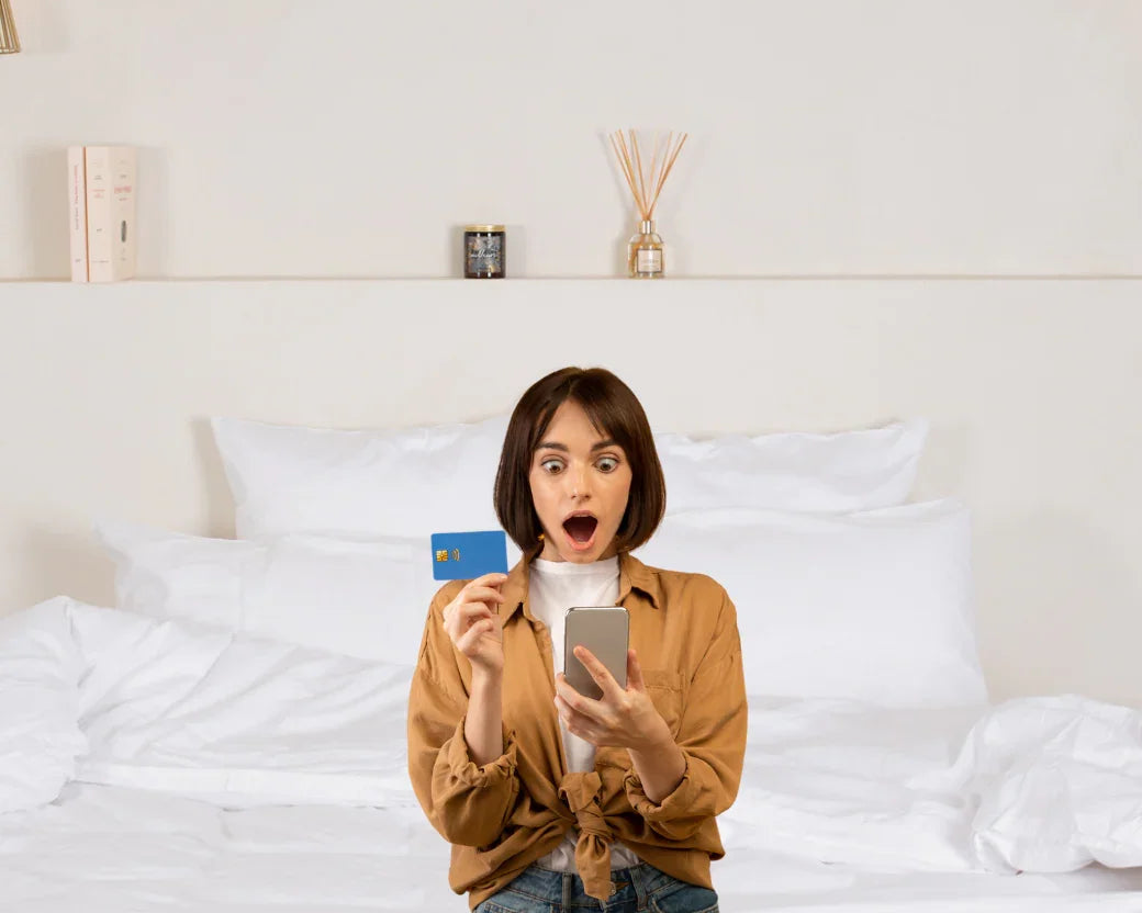 Jeune femme surprise tenant une carte bancaire bleue et un smartphone dans une chambre lumineuse