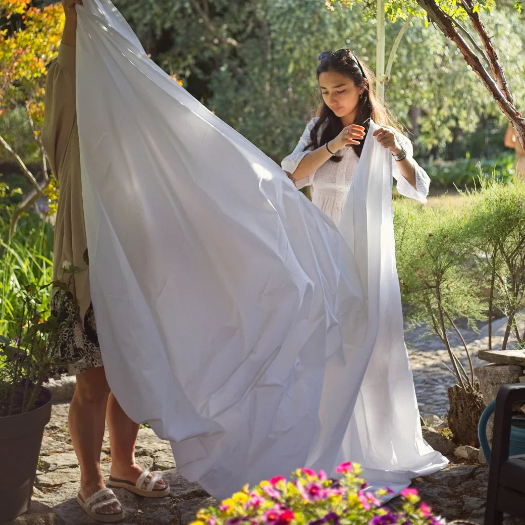 Deux femmes tiennent un grand drap blanc dans un jardin ensoleillé avec des fleurs colorées
