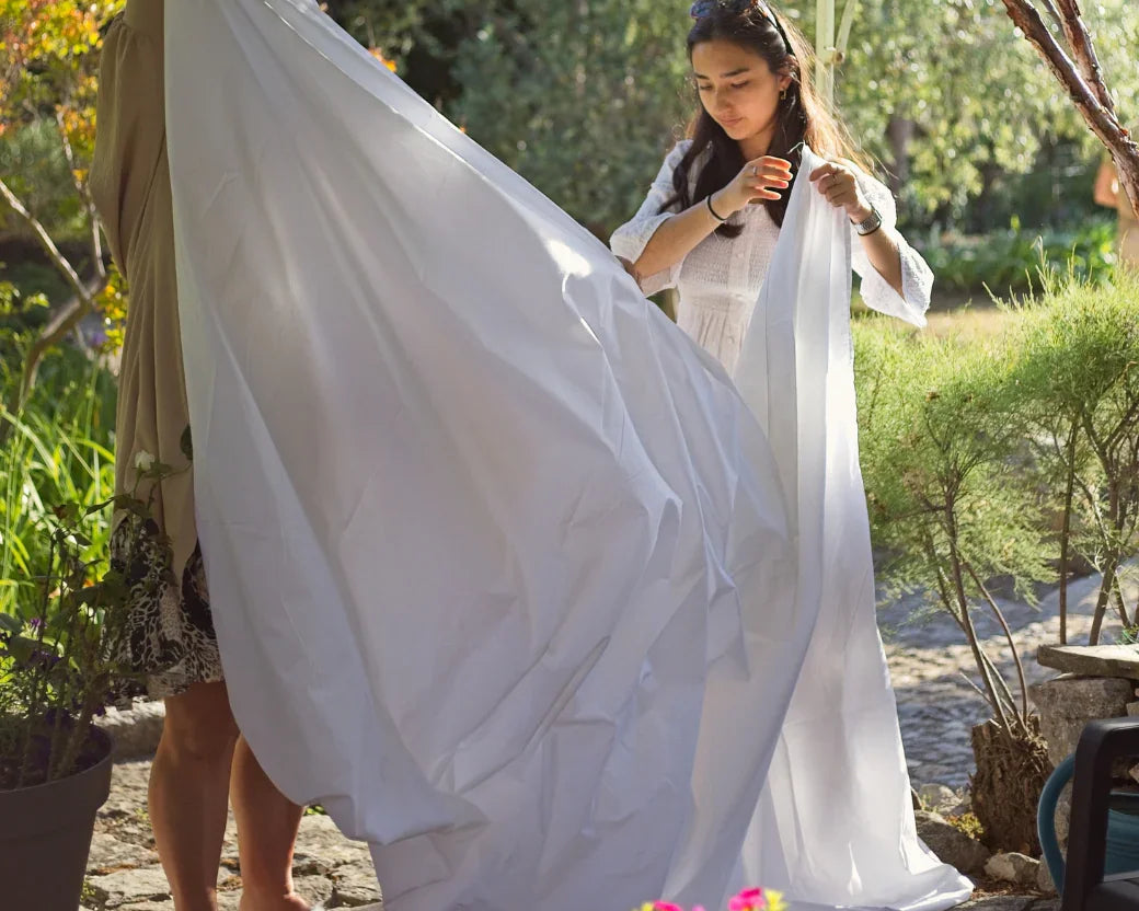Deux femmes tiennent un grand drap blanc dans un jardin ensoleillé avec des fleurs colorées