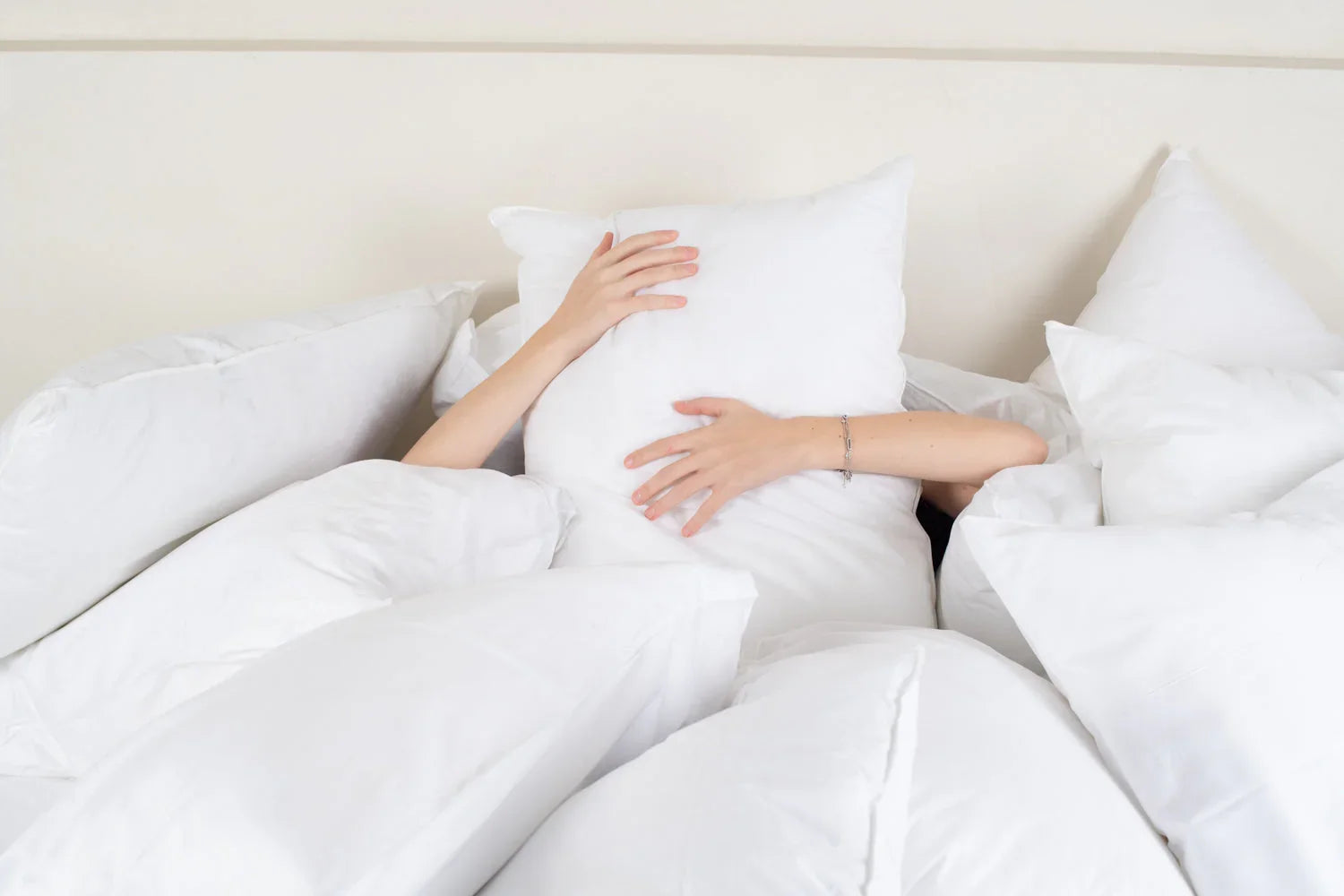 Personne cachée derrière un coussin blanc, entourée de nombreux coussins blancs en lit